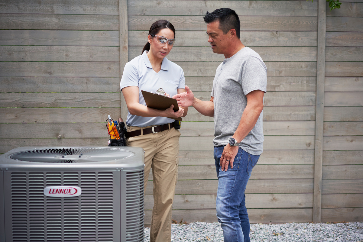 Female HVAC technician discussing service details with a homeowner while referencing a clipboard, standing next to a Lennox outdoor air conditioning unit in a fenced backyard area.