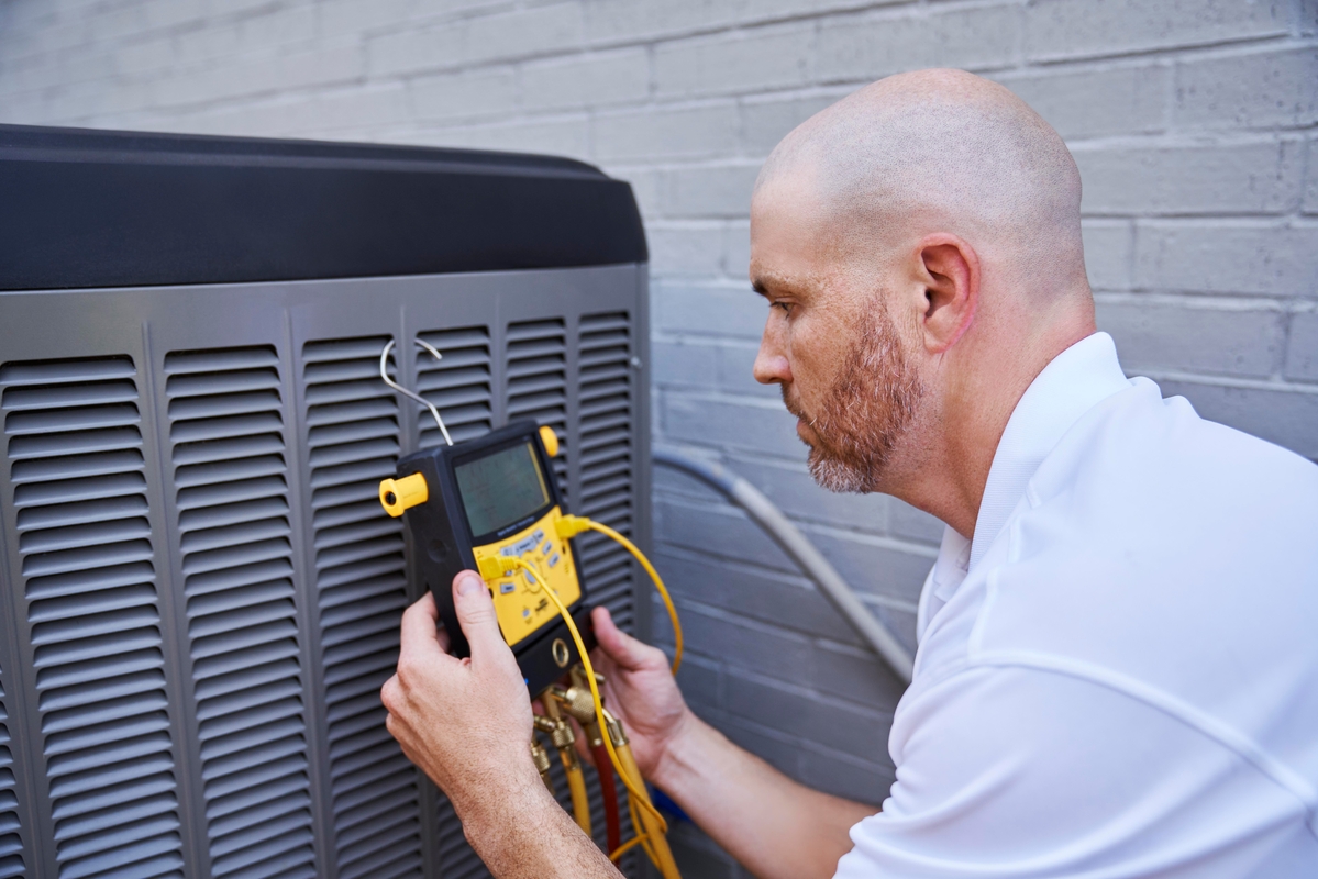 HVAC technician using a digital manifold gauge to check refrigerant levels on a residential outdoor air conditioning unit, positioned next to a gray brick wall.