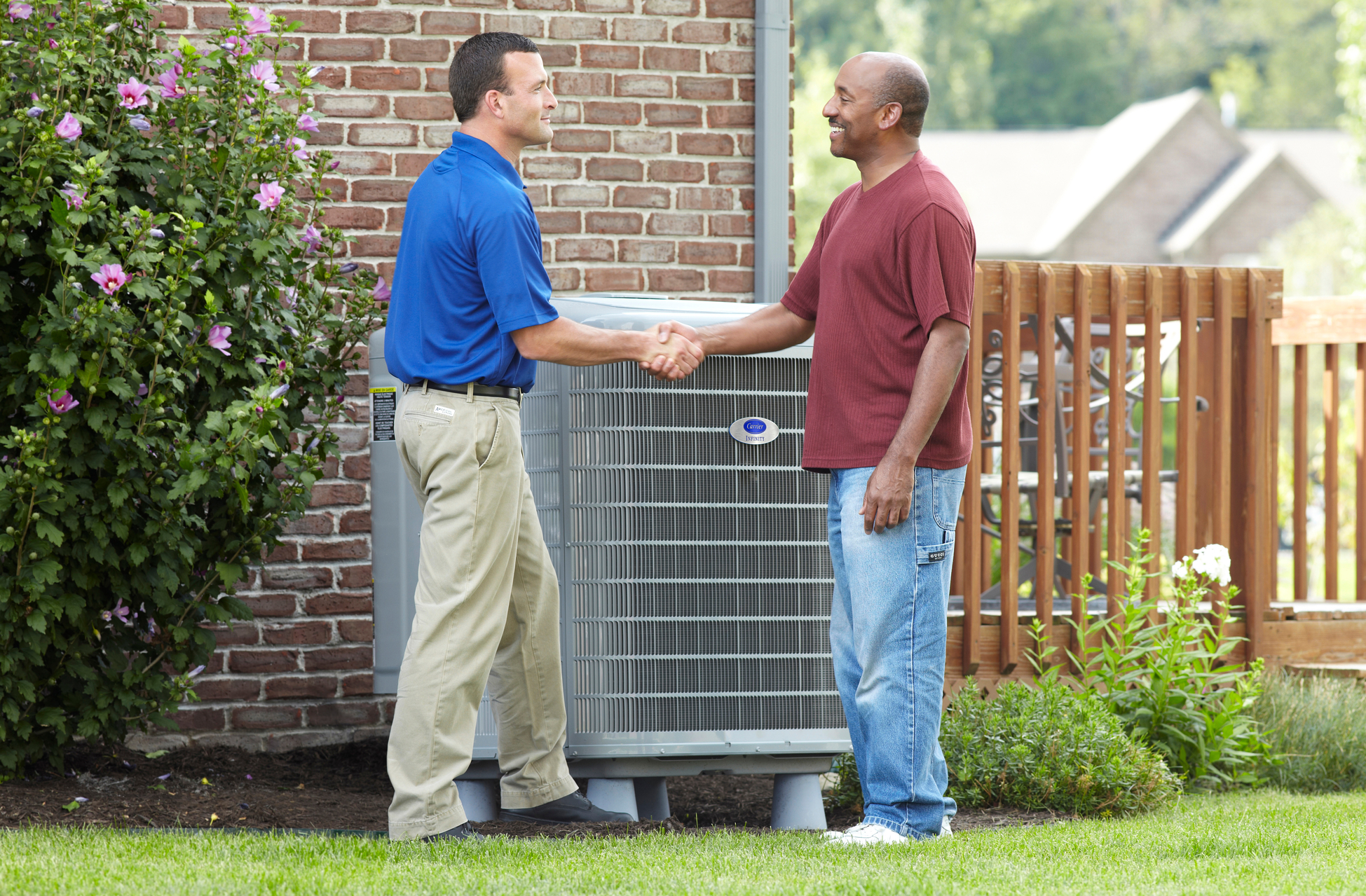 HVAC technician shaking hands with a smiling homeowner in a backyard next to a newly installed residential air conditioning unit, with a brick wall and wooden deck in the background.