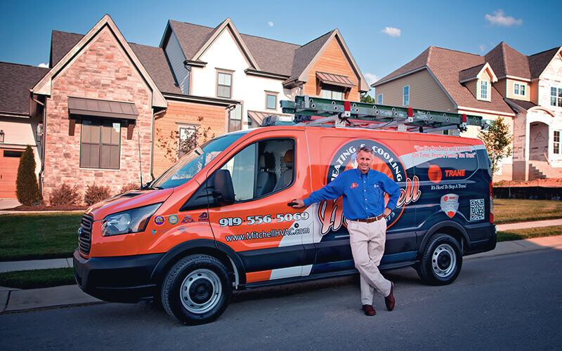 HVAC technician in a blue uniform shirt leaning against a brightly branded service van parked in a suburban neighborhood, with modern homes in the background and ladders secured on the van's roof.
