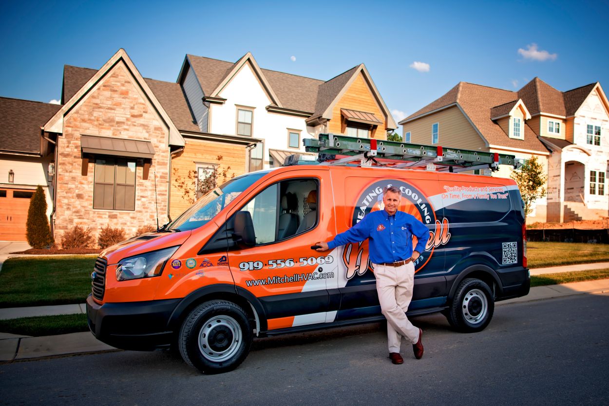HVAC technician in a blue uniform shirt leaning against a brightly branded service van parked in a suburban neighborhood, with modern homes in the background and ladders secured on the van's roof.